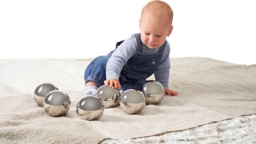 Ein Baby in einem blauen Overall und einem gestreiften Hemd sitzt auf einer beige-weißen Decke und streckt die Hand aus, um eine der sechs glänzenden Metallkugeln zu berühren, die vor ihm liegen.