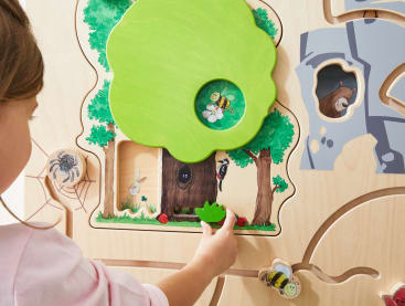 Ein Kind bewegt eine grüne Holzfigur auf einem interaktiven Spielbrett, das einen Baum, Tiere und Illustrationen zum Thema Natur zeigt. Das Brett besteht aus hellem Holz mit ausgeschnittenen Pfaden und gemalten Details.