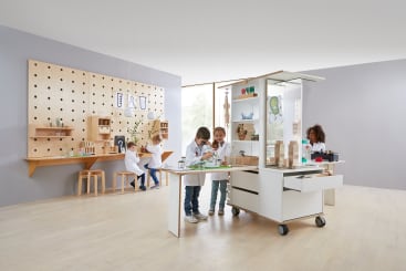 Kinder sind in einem Klassenzimmer mit wissenschaftlichen Aktivitäten beschäftigt. Drei Kinder stehen um einen mobilen Arbeitsplatz mit Laborgeräten, während zwei Kinder an einem an der Wand befestigten Schreibtisch mit Regalen und Hockern im Hintergrund arbeiten.