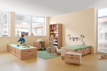 Ein Kind sitzt auf einem niedrigen Holzbett und spielt mit Spielzeug in einem hellen, aufgeräumten Raum mit grünen Matten, Aufbewahrungseinheiten, Regalen, Holzbausteinen und Kunstwerken an der Wand. Große Fenster lassen viel natürliches Licht herein.