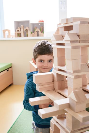 Ein kleines Kind steht in einem Haus, lächelt und schaut in die Kamera, während es mit einem großen, komplexen Bauwerk aus Holzklötzen spielt. Im Hintergrund sind Regale mit weiteren Holzspielzeugen zu sehen.