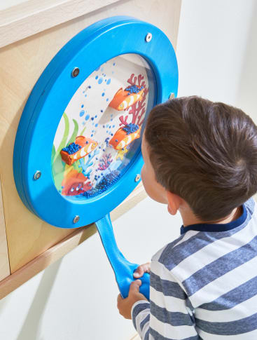 Ein kleines Kind in einem gestreiften Hemd schaut in eine blaue, runde Wandtafel mit bunten Fischen und Unterwasserlandschaften. Das Kind hält einen blauen Griff, der an der Tafel befestigt ist.