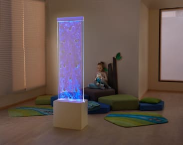 Ein kleines Kind sitzt auf einem gepolsterten Sitz und hält ein Stofftier in einem Raum mit hellem Holzboden, bunten Matten und einer großen rechteckigen LED-Röhre, die im Vordergrund blau leuchtet.