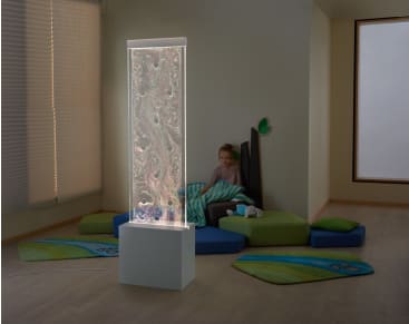 Ein Mädchen sitzt auf bunten Schaumstoffmatten und -kissen in einer Zimmerecke, eingewickelt in eine Decke. Im Vordergrund steht eine hohe, beleuchtete Seifenblasensäule mit wirbelnden Mustern. Der Raum hat einen Holzfußboden und große Fenster.