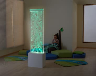 Ein kleines Kind sitzt auf einer gepolsterten Spielfläche in einem sanft beleuchteten Raum und blickt auf eine hohe, beleuchtete Tafel mit einer wirbelnden grünen Flüssigkeit darin. Kissen und Matten mit blauen und grünen Mustern liegen auf dem Holzboden.