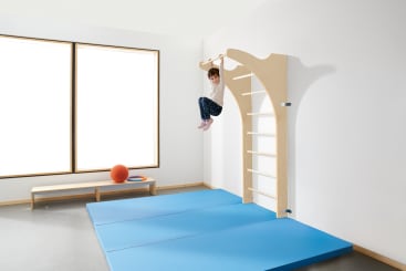 In einem hellen Raum mit großen Fenstern, blauen Turnmatten auf dem Boden und einer niedrigen Bank mit einem orangefarbenen Ball und Ring hängt ein Kind an hölzernen Kletterstangen, die an der Wand befestigt sind.