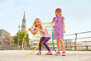 Zwei junge Mädchen stehen draußen auf einer Betonfläche und lächeln. Ein Mädchen in gemusterten Leggings und einem T-Shirt hält einen gelben Hula-Hoop-Reifen, während das andere in einem gestreiften Kleid und einer Mütze einen grünen Hula-Hoop-Reifen hält. Im Hintergrund sind Gebäude zu sehen.