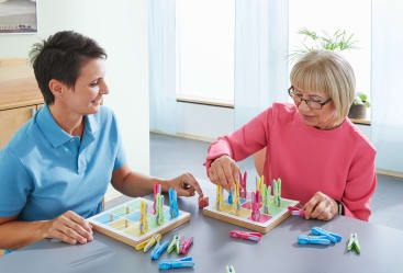Zwei Frauen sitzen an einem Tisch und spielen ein Spiel mit bunten Wäscheklammern und Holzbrettern mit quadratischem Querschnitt. Eine Frau setzt eine Wäscheklammer auf ihr Brett, während die andere lächelnd zusieht.