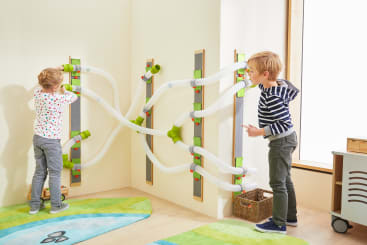 Zwei Kinder spielen mit einem an der Wand befestigten Röhrensystem in einem hellen Raum. Die Rohre sind an den Wänden befestigt und in der Ecke miteinander verbunden, so dass Gegenstände durch sie hindurchfallen oder geblasen werden können. Der Boden ist mit bunten Teppichen ausgelegt.