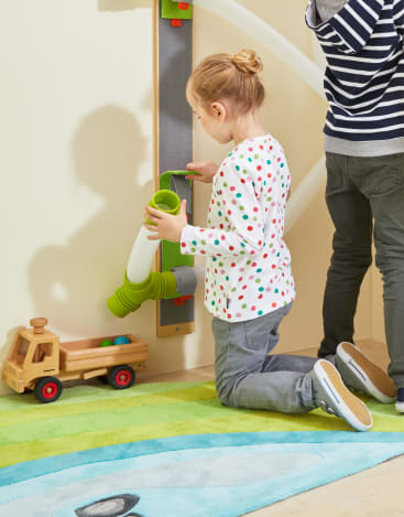 Ein Kind kniet auf einem grün-blauen Teppich und stellt eine grün-weiße Plastikrutsche auf ein an der Wand befestigtes vertikales Spielgerät, in dessen Nähe sich ein hölzerner Spielzeuglaster befindet. Ein anderes Kind steht daneben und schaut weg.