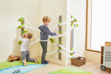 Zwei kleine Kinder spielen mit einer Murmelbahn, die in einem hellen Raum an der Wand befestigt ist. Der Junge stellt die Bahn ein, während das Mädchen auf dem Boden kniet und mit dem unteren Teil interagiert. Ein Spielzeug-LKW und ein Korb stehen in der Nähe.