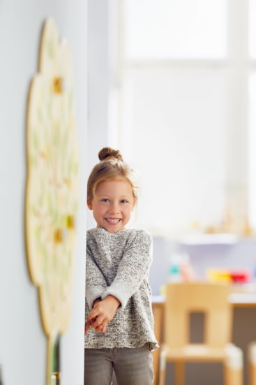 Ein junges Kind mit hellbraunem Haar in einem Dutt steht teilweise hinter einer Wand und lächelt. Der Hintergrund zeigt ein Klassenzimmer mit einem Holzstuhl und unscharfen Unterrichtsmaterialien.