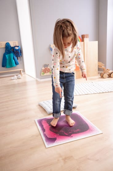 Ein kleines Kind steht barfuß auf einer lila und schwarz gefärbten sensorischen Gelmatte, drückt mit einem Fuß darauf und zeigt nach unten. Das Kind befindet sich in einem Spielzimmer mit hellem Holzfußboden und Spielzeug im Hintergrund.