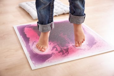 Ein Kind steht barfuß auf einer quadratischen Sinnesmatte mit einem rosa und lila Muster. Die Jeans des Kindes sind hochgekrempelt, und die Matte liegt auf einem hellen Holzboden. Im Hintergrund befindet sich ein weißes Kissen.