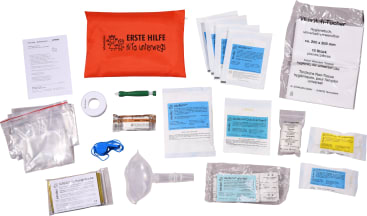 Ein Erste-Hilfe-Kasten mit einem roten Beutel mit der Aufschrift ERSTE HILFE, der auf einer weißen Fläche Verbandsmaterial, Tücher, Mull, medizinisches Klebeband, Scheren, Handschuhe, eine Gesichtsmaske, eine Trillerpfeife und Informationsbroschüren enthält.