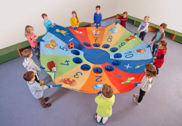 Zwölf Kinder stehen im Kreis und halten einen bunten, nummerierten Fallschirm mit Tiermotiven und Löchern in der Hand, auf dem ein rot-schwarzer Ball ruht. Der Raum hat einen grauen Boden und weiße Wände.