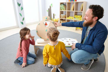 Ein Erwachsener und zwei kleine Kinder sitzen in einem Klassenzimmer auf dem Boden. Der Erwachsene zeigt auf ein hölzernes Spinnrad mit Tierbildern, während ein Kind beobachtet und ein anderes ein Buch mit Tierbildern anschaut.