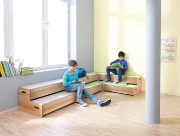 Zwei Jungen sitzen auf modularen Holzbänken mit grauen Polstern in einem hellen Eckzimmer. Ein Junge schreibt in ein Notizbuch, während der andere ein Buch liest. Im Hintergrund sind Bücher und Lernspielzeug zu sehen.