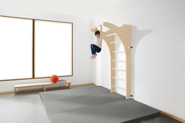In einem hellen Raum mit großen Fenstern, einer Bank, einem orangefarbenen Ball und grauen gepolsterten Matten auf dem Boden hängt ein Kind an einer horizontalen Holzstange, die an einem an der Wand befestigten Klettergerüst angebracht ist.