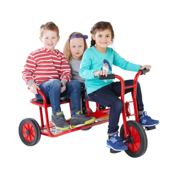 Drei Kinder fahren auf einem roten Tandem-Dreirad. Zwei Kinder sitzen nebeneinander auf dem Rücksitz, während ein Kind vorne sitzt und lenkt. Alle tragen Freizeitkleidung und Turnschuhe. Der Hintergrund ist einfarbig weiß.
