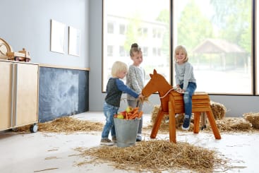 Drei kleine Kinder spielen drinnen mit Heu, einem Holzpferd und einem mit Äpfeln und Karotten gefüllten Metalleimer. Große Fenster geben den Blick frei auf eine helle Szene im Freien im Hintergrund.