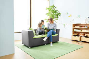 Ein Erwachsener und zwei Kinder sitzen auf einem grau-grünen Sofa und lesen gemeinsam ein Buch in einem hellen Raum mit einem großen Fenster, einem grünen Teppich, einer Zimmerpflanze und Kinderspielzeug und -büchern in Regalen.
