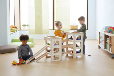 Drei kleine Kinder spielen mit Spielzeugautos und einer hölzernen Rampenstruktur in einem hell erleuchteten Klassenzimmer mit hellen Möbeln, einem grünen Kissen und Regalen mit Spielzeug im Hintergrund.