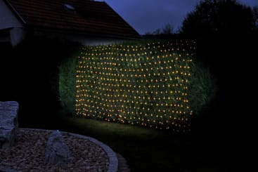 Eine rechteckige Hecke ist mit einem Gitter aus gleichmäßig verteilten, kleinen gelben Lichtern verziert, die in der Nacht leuchten. Im Vordergrund sind ein Teil eines weißen Hauses und ein Steingarten zu sehen.
