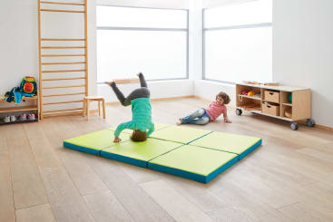 Zwei kleine Kinder spielen in einem hellen Raum mit Holzboden. Ein Kind macht einen Handstand auf einer grünen Matte, während das andere in der Nähe auf dem Boden sitzt. Im Hintergrund sind Regale und Spielzeug zu sehen.