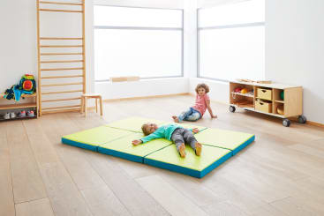 Zwei Kinder befinden sich in einem hellen, minimalistischen Raum mit Holzboden. Ein Kind liegt auf einer grün-blauen Turnmatte, während das andere in der Nähe sitzt. Im Hintergrund befinden sich ein hölzernes Klettergerüst und Regale mit Spielzeug.