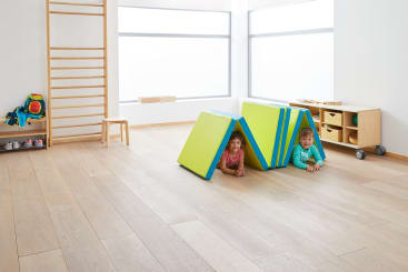 In einem hellen, minimalistischen Spielzimmer mit Holzböden und Turngeräten liegen zwei Kinder auf dem Boden unter einer dachartigen Struktur aus gefalteten Schaumstoffmatten.