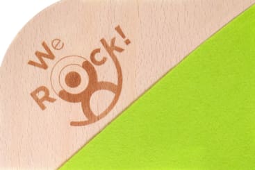 Eine helle Holzoberfläche mit dem eingravierten Schriftzug "We Rock!" und einer stilisierten menschlichen Figur, die teilweise von einem diagonalen Stück hellgrünen Filzes bedeckt ist.