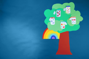 Ein Papierbaum mit grünen Blättern und einem roten Stamm steht vor einem blauen Hintergrund. An dem Baum hängen fünf Papiergläser mit Zeichnungen von Obst und Gemüse. Hinter dem Baumstamm ist teilweise ein Regenbogen zu sehen.