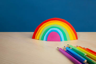 Ein ausgeschnittener Regenbogen aus Papier steht auf einer hellen Holzfläche vor einem blauen Hintergrund. Auf dem Tisch rechts vom Regenbogen liegen mehrere Buntstifte, die in Regenbogenreihenfolge angeordnet sind.