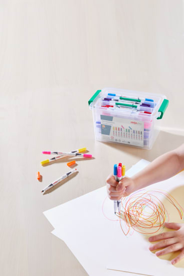 Die Hand eines Kindes zeichnet mit Markern bunte Spiralen auf weißes Papier. Mehrere bunte Stifte und Textmarker liegen in der Nähe verstreut, und eine durchsichtige Schachtel mit Stiften steht im Hintergrund auf einem hellen Holztisch.