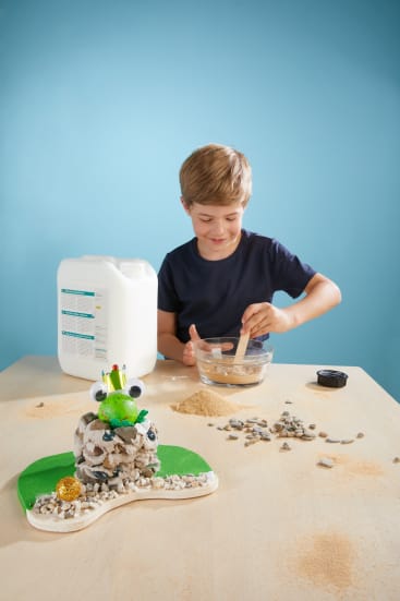 Ein Junge mischt Zutaten in einer Schüssel an einem Tisch, daneben stehen ein Krug mit Flüssigkeit und kleine Behälter. Auf dem Tisch liegen verstreut Steine, Sand und ein froschförmiges Kunstwerk aus Steinen und grünem Material. Blauer Hintergrund.