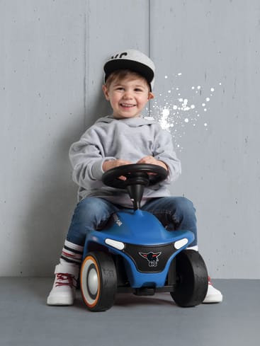 Ein kleines Kind, das einen grauen Kapuzenpullover, blaue Jeans und eine Baseballmütze trägt, sitzt auf einem blauen Spielzeugauto vor einer grauen Wand. Das Kind lächelt und hält das Lenkrad fest.