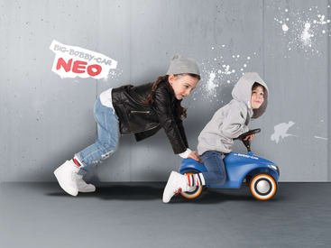 Zwei Kinder spielen in einem Haus vor einer grauen Wand. Ein Kind in einem grauen Kapuzenpulli fährt ein kleines blaues Spielzeugauto, während ein anderes Kind in einer Lederjacke und einer grauen Mütze von hinten schiebt. Die Aufschrift BIG-BOBBY-CAR NEO steht an der Wand.
