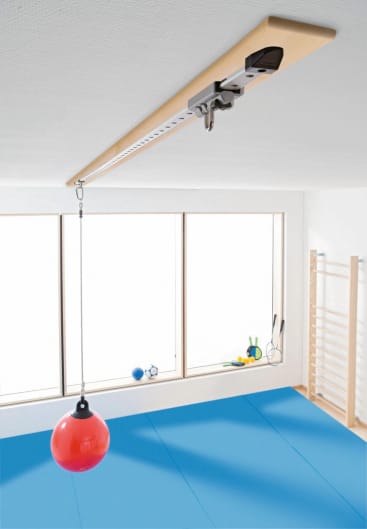 In einem hellen Raum mit blauen Fußmatten und großen Fenstern hängt ein roter Ball an einer Schnur, die an einem an der Decke montierten Schienensystem befestigt ist, von der Decke herab.