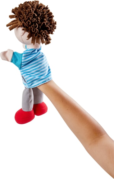 Ein Personenarm trägt eine Handpuppe aus Stoff, die wie ein Kind mit braunem Garnhaar, einem blau-weiß gestreiften Hemd, einer grauen Hose und roten Schuhen aussehen soll. Die Puppe ist nach links gerichtet.