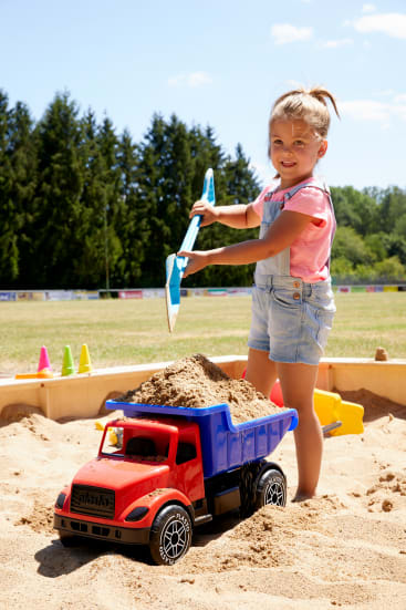 Ein kleines Kind steht in einem Sandkasten, lächelt und hält eine mit Sand gefüllte Schaufel über einen großen Spielzeug-Kipplaster. Der Sandkasten steht im Freien mit Bäumen und einer Wiese im Hintergrund.