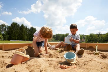Zwei kleine Kinder spielen in einem Sandkasten unter einem blauen Himmel mit Wolken. Das Mädchen gräbt im Sand, während der Junge den Sand mit einem Eimer schöpft. Spielzeug und Sandformen sind um sie herum verstreut. Im Hintergrund sind Bäume und Gras zu sehen.