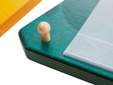 Nahaufnahme einer Ecke eines grün gefärbten Holzbretts mit einem kleinen zylindrischen Holzpflock und einer transparenten Plastikfolie, die an der Oberfläche befestigt ist. Im Hintergrund ist ein Teil eines gelben Holzbretts zu sehen.