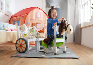 Ein kleines Kind sitzt auf einem Spielzeugpferd in einem hell erleuchteten Schlafzimmer, lächelt und spielt. In einem Spielzeugwagen in der Nähe sitzen Plüschtiere, und im Hintergrund ist ein rotes Spielzelt aufgebaut.