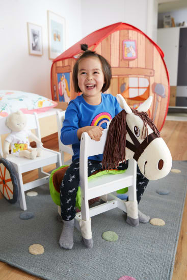 Ein kleines Kind sitzt auf einem Spielzeugpferd in einem Spielzimmer und lächelt. Dahinter stehen ein kleines Zelt in Form eines Hauses und ein weißer Stuhl, auf dem ein Stofftier sitzt. Das Zimmer hat einen gemusterten Teppich und gerahmte Bilder an der Wand.