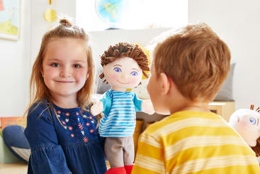 Ein junges Mädchen in einem blauen Kleid hält eine Puppe mit braunem Garnhaar und einem gestreiften Hemd. Ein kleiner Junge in einem gelben Hemd steht ihr gegenüber. Eine Puppe mit orangefarbenem Haar ist teilweise rechts zu sehen. Der Schauplatz ist ein heller Raum.
