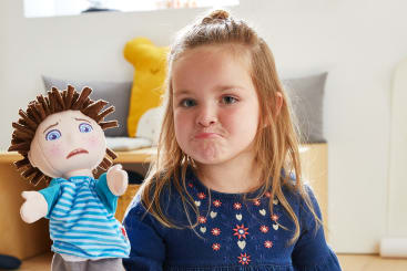 Ein junges Mädchen in einem blauen Kleid hält eine Handpuppe mit einem besorgten Gesichtsausdruck. Das Mädchen ahmt den Ausdruck der Puppe nach, indem es die Lippen schürzt. Der Hintergrund zeigt ein helles Zimmer mit Spielzeug und Kissen.