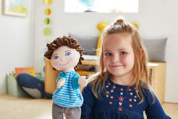 Ein junges Mädchen mit blondem Haar und einem blauen Kleid hält eine Puppe mit braunem Garnhaar, blauen Augen und einem gestreiften Hemd in einem hell erleuchteten Raum mit Spielzeug und Büchern im Hintergrund.