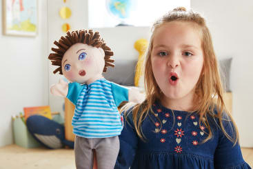 Ein junges Mädchen in einem blauen Kleid hält eine Handpuppe mit braunem Garnhaar und einem gestreiften Hemd. Der Mund des Mädchens ist geöffnet, als ob es sprechen oder singen würde. Die Szene spielt sich in einem hell erleuchteten Raum ab.