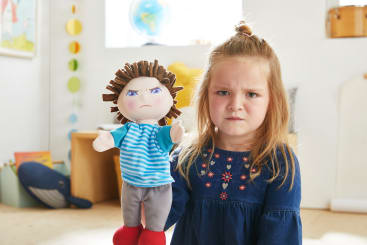 Ein junges Mädchen mit einem ernsten Gesichtsausdruck hält eine Handpuppe mit einem wütenden Gesicht. Sie steht in einem hellen Raum, in dem im Hintergrund Kinderspielzeug und Bücher zu sehen sind.